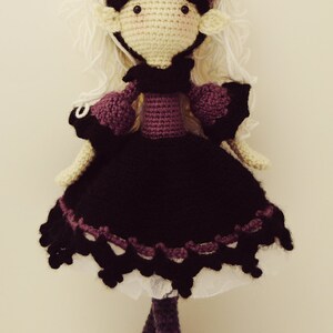 May include: A crocheted doll with white hair, a purple dress, and black shoes. The doll is wearing a black hat with a purple flower.