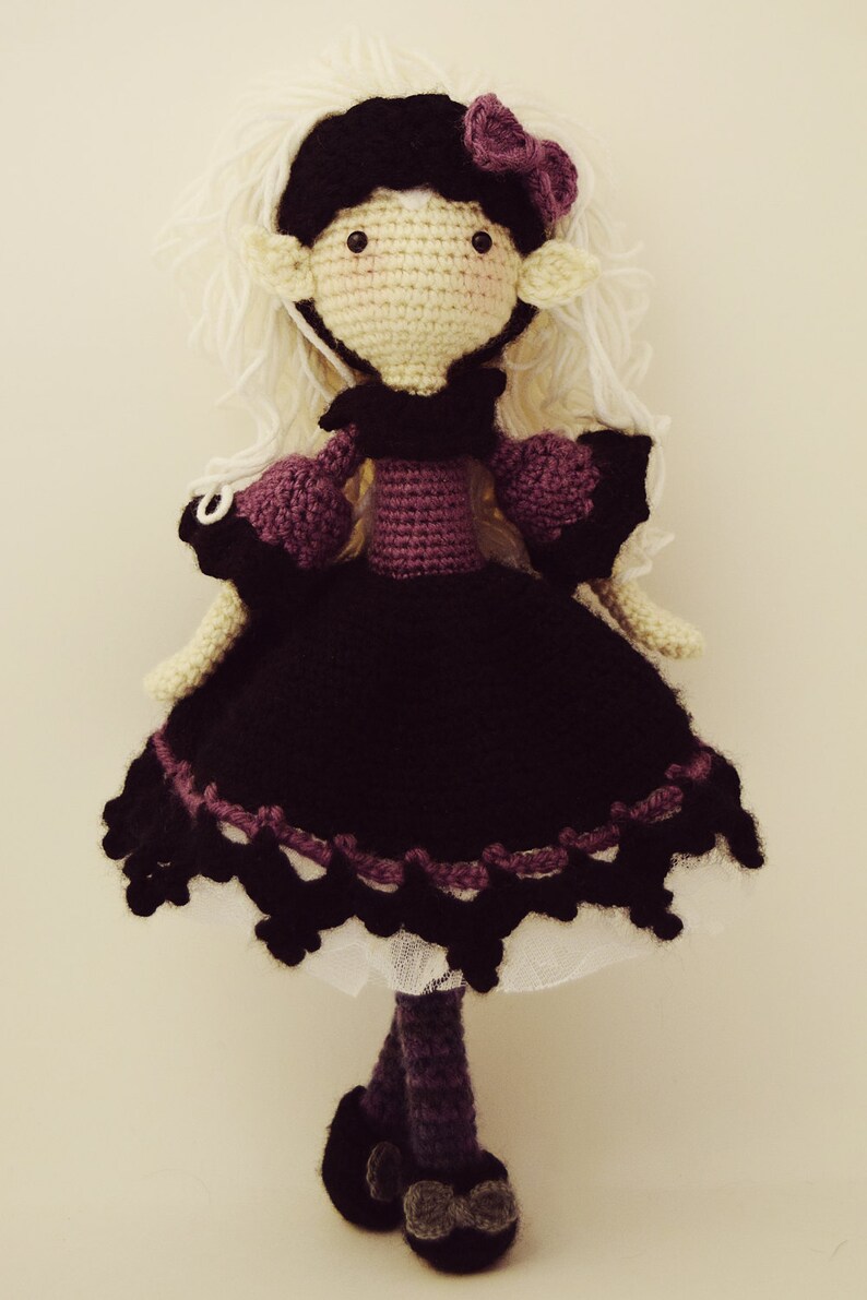 May include: A crocheted doll with white hair, a purple dress, and black shoes. The doll is wearing a black hat with a purple flower.