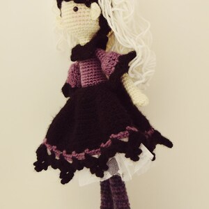 May include: Crocheted doll with long white yarn hair, wearing a purple and black dress with a white lace trim and black shoes.