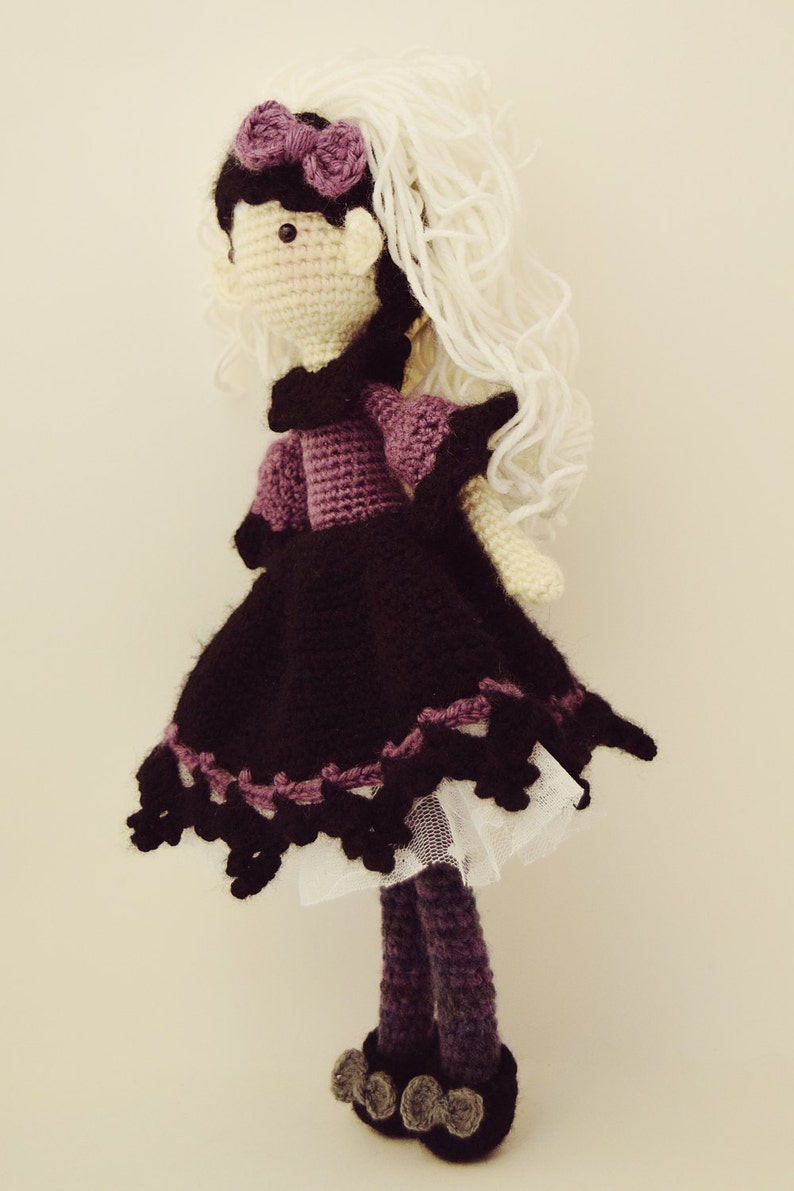 May include: Crocheted doll with long white yarn hair, wearing a purple and black dress with a white lace trim and black shoes.