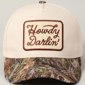 May include: A cream-coloured baseball cap with a camouflage brim. The cap features a rectangular patch with the embroidered words "Howdy Darlin'" in brown script. The top button is also camouflage.