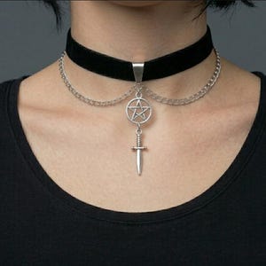 May include: Black velvet choker necklace with a silver-tone pentagram pendant and a small dagger charm. The necklace features a chain detail and is worn around the neck. The overall style is gothic and edgy.