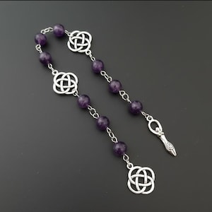 Könnte beinhalten: Ein silberfarbenes Armband im Rosenkranzstil mit Amethystperlen und keltischen Knotenanhängern. Das Armband hat einen Anhänger einer stilisierten weiblichen Figur. Die Perlen sind tiefviolett und das Metall glänzend.