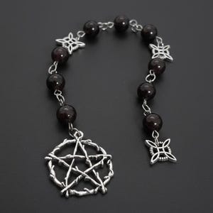 Può includere: Una collana argentata con perline rotonde rosso scuro e ciondoli decorativi. I ciondoli includono un pentagramma e forme floreali stilizzate. La collana è mostrata su uno sfondo scuro.