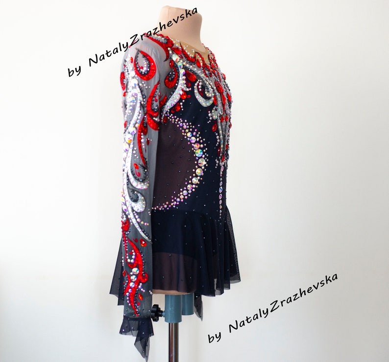 May include: A black and red figure skating dress with intricate rhinestone and sequin embellishments. The long-sleeved dress features a sheer mesh overlay on the sleeves and upper bodice, with a flared skirt. The design includes a crescent of rhinestones on the front.