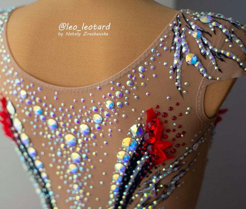 May include: A tan leotard with intricate embellishments, including iridescent rhinestones and red floral accents. Black and silver painted details create a striking contrast. The text "@leo_leotard by Nataly Zrazhevsk" is visible.