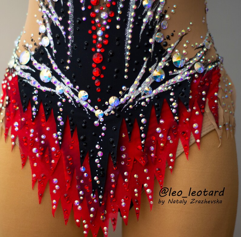 May include: A close-up of a black and red leotard with intricate rhinestone and sequin detailing. The leotard features a high neckline and is embellished with a variety of iridescent and red rhinestones. The design includes a jagged red skirt.