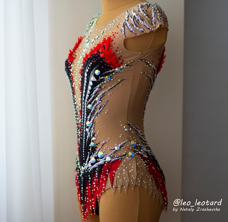 May include: A rhythmic gymnastics leotard on a mannequin. The leotard features a nude-colored base with black, red, and white accents. It is embellished with numerous iridescent rhinestones. The design includes feather-like patterns and a high neckline. The text "@leo_leotard" is visible.