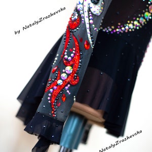 May include: Black figure skating dress with intricate red and crystal embellishments. The sleeve features a vibrant red design with sparkling crystals and iridescent accents. The dress has a sheer skirt and is adorned with additional crystals.