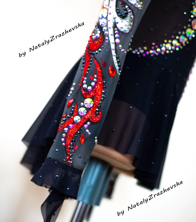 May include: Black figure skating dress with intricate red and crystal embellishments. The sleeve features a vibrant red design with sparkling crystals and iridescent accents. The dress has a sheer skirt and is adorned with additional crystals.