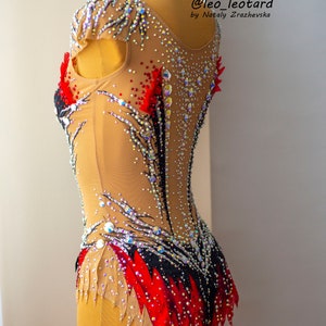 May include: A tan and black leotard with red accents and sparkling rhinestones. The leotard has a high neckline and is decorated with intricate patterns. The text "@leo_leotard by Nataly Zrazhevska" is visible.