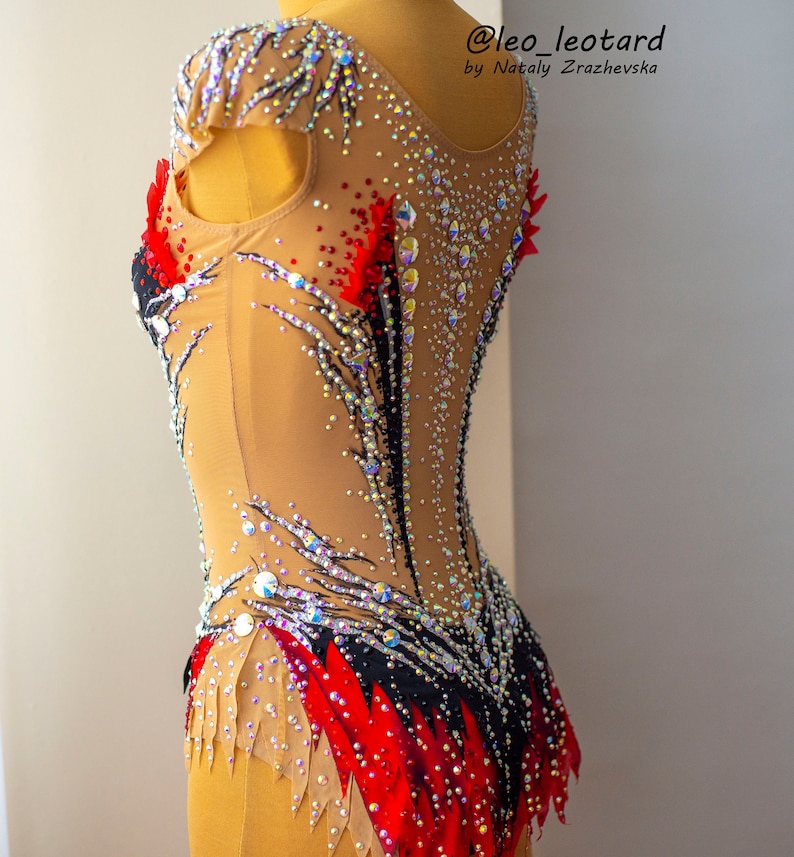 May include: A tan and black leotard with red accents and sparkling rhinestones. The leotard has a high neckline and is decorated with intricate patterns. The text "@leo_leotard by Nataly Zrazhevska" is visible.