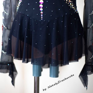 May include: A black figure skating dress with a flared skirt and long sleeves. The bodice and skirt are embellished with sparkling rhinestones and iridescent crystals. The sleeves have a sheer, ruffled design. The dress is displayed on a mannequin.