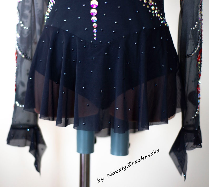 May include: A black figure skating dress with a flared skirt and long sleeves. The bodice and skirt are embellished with sparkling rhinestones and iridescent crystals. The sleeves have a sheer, ruffled design. The dress is displayed on a mannequin.