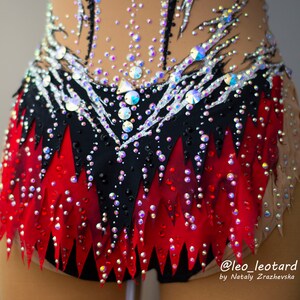 May include: A close-up of a rhythmic gymnastics leotard. The leotard features a black and red skirt design with a jagged edge, adorned with sparkling rhinestones. The top is nude-colored with black and silver accents and more rhinestones.