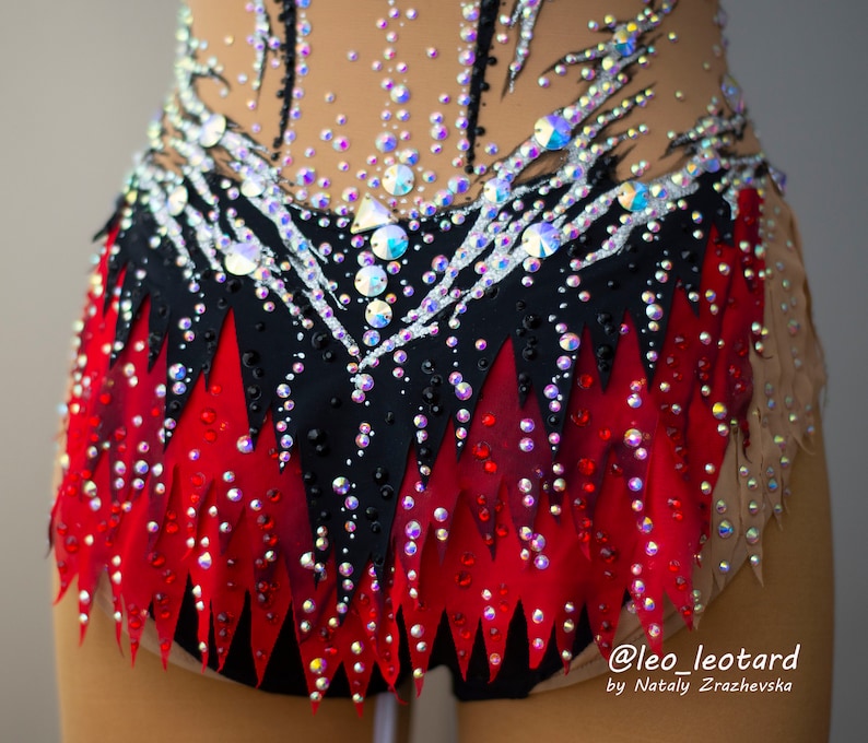 May include: A close-up of a rhythmic gymnastics leotard. The leotard features a black and red skirt design with a jagged edge, adorned with sparkling rhinestones. The top is nude-colored with black and silver accents and more rhinestones.