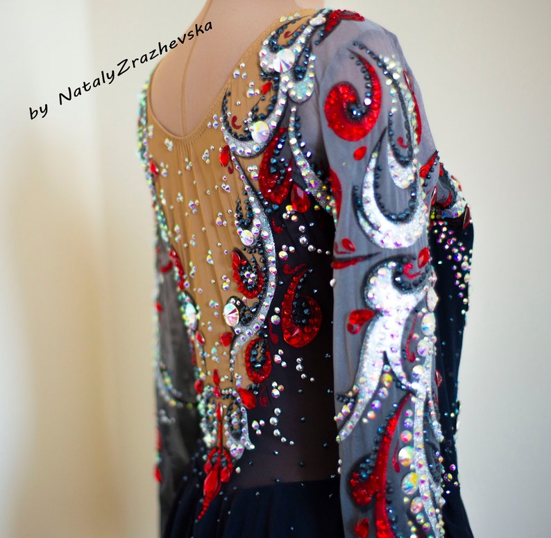 May include: A long-sleeved leotard with intricate embellishments. The garment features a sheer nude mesh back and sleeves, adorned with red, silver, and clear crystals. The design includes swirling patterns and a black skirt. The text "by Nataly Zrazhevska" is visible.