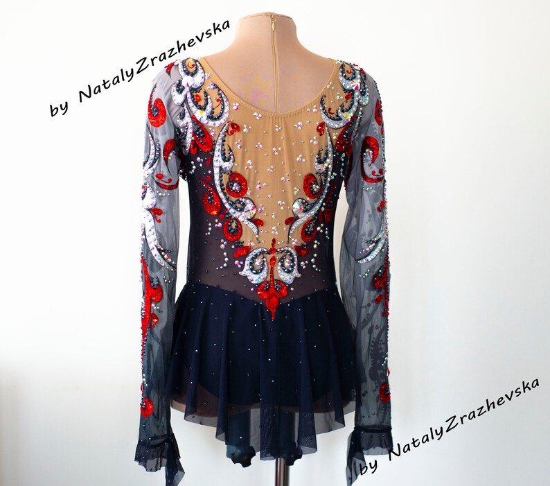 May include: A figure skating dress with long sleeves. The bodice is beige with intricate red, black, and silver embellishments. The sleeves are sheer gray, and the skirt is navy blue with a ruffled hem. The text "by Nataly Zrazhevska" is visible.