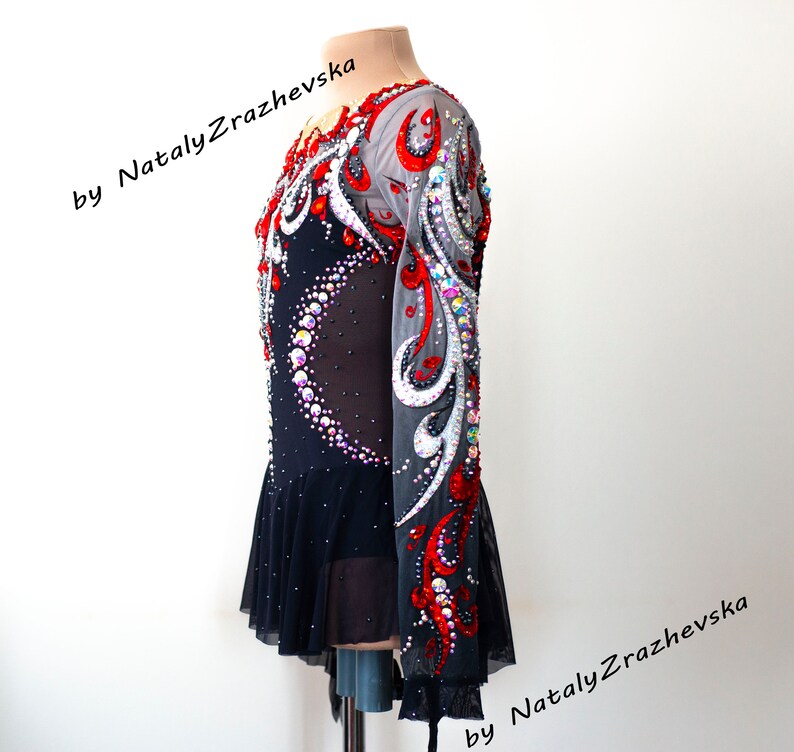 May include: A black and red figure skating dress with long sleeves, embellished with rhinestones and decorative patterns. The dress features a sheer mesh overlay and a ruffled skirt. Text on the image reads "by Nataly Zrazhevsk".