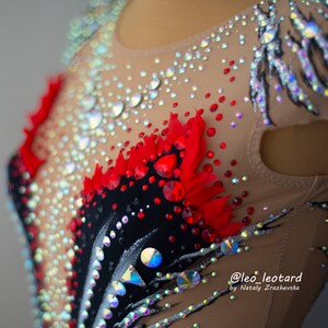 May include: Close-up of a beige leotard with intricate embellishments. The leotard features a black and red design with sparkling rhinestones and crystals. The design includes a feather-like pattern and is adorned with various sizes of iridescent gems.