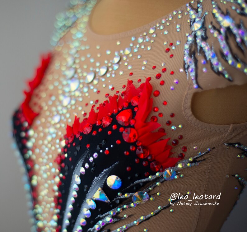May include: Close-up of a beige leotard with intricate embellishments. The leotard features a black and red design with sparkling rhinestones and crystals. The design includes a feather-like pattern and is adorned with various sizes of iridescent gems.