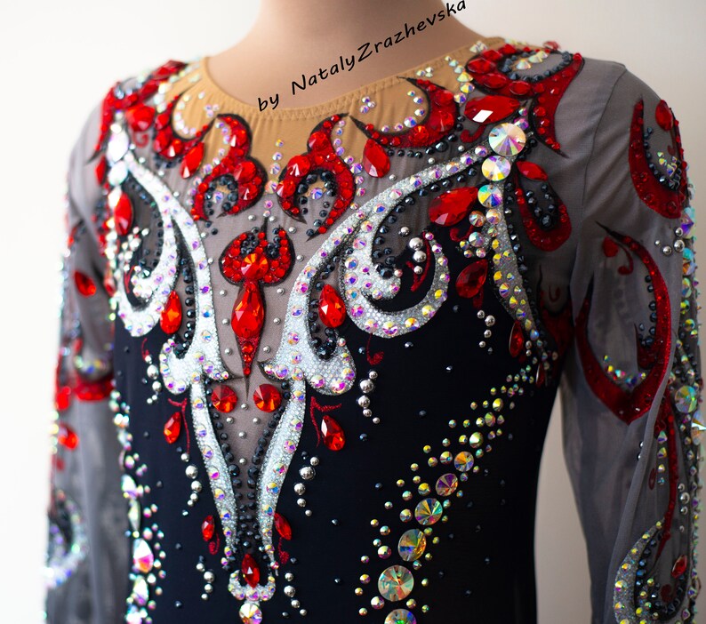 May include: A black leotard with long sheer sleeves, embellished with red and iridescent rhinestones. The design features a central pattern of silver and red accents, with the text "by Nataly Zrazhevsk" visible.