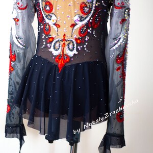 May include: A black and nude leotard with long, sheer sleeves. The bodice is embellished with red, white, and clear rhinestones in an ornate design. The skirt is a dark navy blue with scattered rhinestones. The text "by Nataly Zrazhevska" is visible.