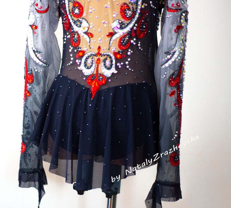 May include: A black and nude leotard with long, sheer sleeves. The bodice is embellished with red, white, and clear rhinestones in an ornate design. The skirt is a dark navy blue with scattered rhinestones. The text "by Nataly Zrazhevska" is visible.