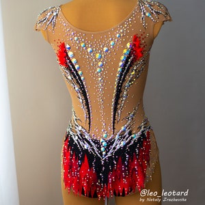 May include: A tan-colored leotard with intricate embellishments. The garment features a black and red design with sparkling rhinestones and crystals. The leotard is designed for gymnastics or dance performances.