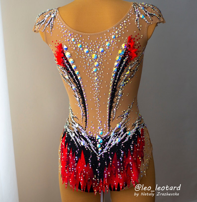 May include: A tan-colored leotard with intricate embellishments. The garment features a black and red design with sparkling rhinestones and crystals. The leotard is designed for gymnastics or dance performances.