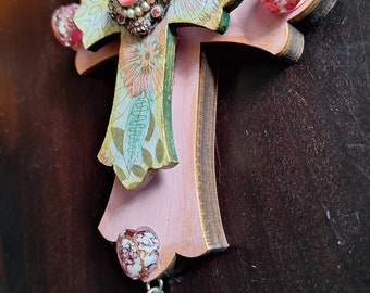 May include: A pink wooden cross with a floral design and a jeweled embellishment. The cross has a light blue glass bead hanging from the bottom.