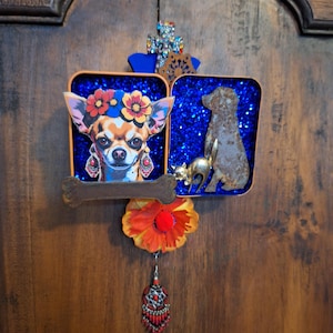 May include: A decorative wall hanging featuring a chihuahua with floral headwear, a metal dog figurine, and a small gold cat. The artwork includes a cross, a bone-shaped accent, and a dangling red and silver ornament. The background is a wooden surface.