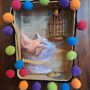 Puede incluir: Una bandeja decorativa con un retrato de gato, una jaula de pájaros en miniatura y una figura de ratón dorada. La bandeja está adornada con una guirnalda de pompones coloridos en tonos rosa, azul, verde, naranja y morado. El retrato del gato tiene un collar dorado.
