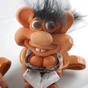Può includere: Una statuetta in ceramica di un troll con un'acconciatura di capelli neri e pelosi, che indossa un gilet a motivi marroni e neri, tenendo un libro tra le mani. Il troll ha un ampio sorriso e occhi grandi e rotondi.
