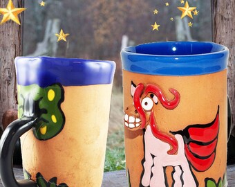 Puede incluir: Dos tazas de cerámica con diseños pintados a mano. Una taza presenta un diseño de mariposa verde y amarilla, mientras que la otra taza presenta un unicornio rosa con alas rojas.