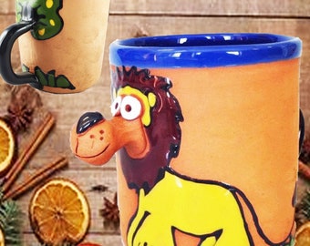 Puede incluir: Una taza de cerámica con un borde azul y una ilustración pintada a mano de un león con un cuerpo amarillo y una melena marrón. La taza tiene un fondo naranja rústico.