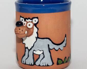 Puede incluir: Una taza de terracota con un borde azul presenta un lobo de dibujos animados pintado a mano con pelaje blanco y una sonrisa de dientes. El lobo está de pie sobre un fondo de hierba verde.