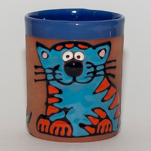 Könnte beinhalten: Eine Terrakotta-Tasse mit blauem Rand und einem blauen und orangefarbenen Comic-Katzen-Design. Die Katze hat große Augen und einen freundlichen Ausdruck.