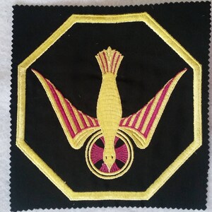 May include: A gold embroidered dove with outstretched wings and a red cross inside a circle, on a black fabric background.