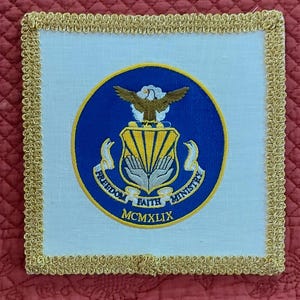 May include: White square patch with a gold border. The patch features a blue circle with a yellow eagle, a shield with yellow stripes, and the words "Freedom, Faith, Ministry" around the shield. The year "MCMXLIX" is printed below the shield.