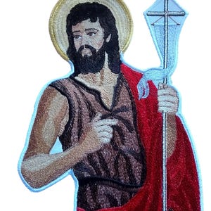 May include: Embroidered patch depicting a religious figure with a halo. The figure has a dark beard, long hair, and is wearing a brown tunic and a red cloak. He holds a staff with a cross. The patch has a blue border and is likely for clothing or accessories.