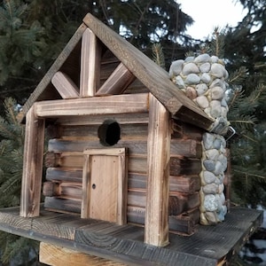 May include: A handcrafted wooden birdhouse in the style of a log cabin, with a stone chimney. The birdhouse has a small door and a circular entrance. Constructed from weathered wood, it rests on a wooden base, suitable for a garden or outdoor space.