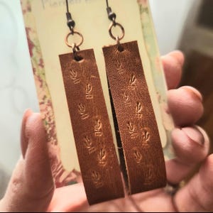 May include: A pair of brown leather earrings with a rectangular shape. Each earring features a series of embossed, stylized flame designs. The earrings hang from copper-colored ear wires.