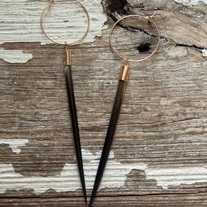 May include: A pair of gold hoop earrings with dark, pointed feather-like pendants. The earrings hang from a gold-colored metal and are set against a weathered, light-colored wooden surface.