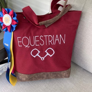 May include: A red canvas tote bag with brown faux leather trim and handles. The bag has the word "EQUESTRIAN" embroidered in white with a pair of stirrups below it.