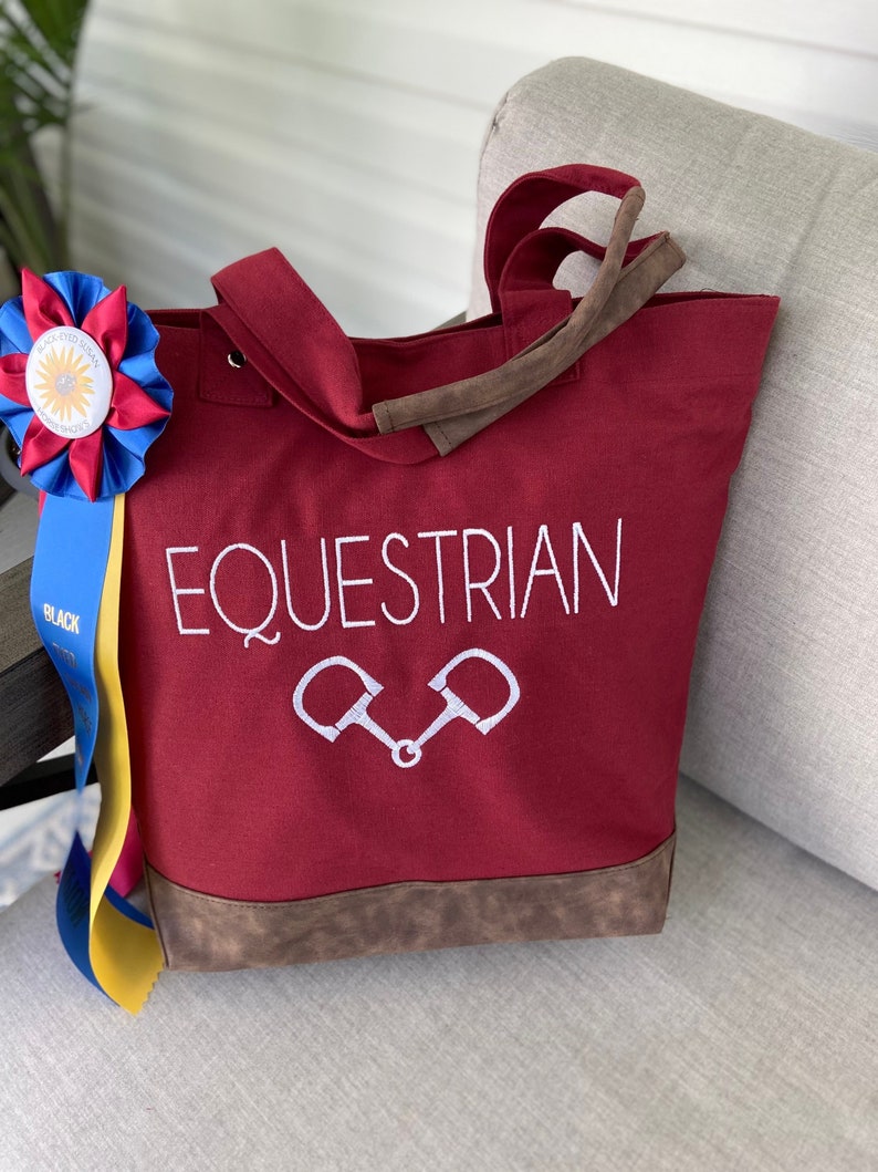 May include: A red canvas tote bag with brown faux leather trim and handles. The bag has the word "EQUESTRIAN" embroidered in white with a pair of stirrups below it.
