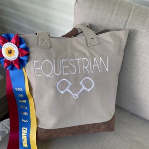 May include: A beige canvas tote bag with brown leather trim. The bag has the word "EQUESTRIAN" embroidered on the front in white, along with a pair of white stirrups. The bag is sitting on a light brown couch.