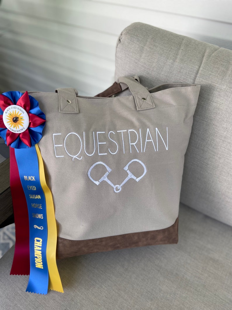 May include: A beige canvas tote bag with brown leather trim. The bag has the word "EQUESTRIAN" embroidered on the front in white, along with a pair of white stirrups. The bag is sitting on a light brown couch.
