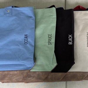 May include: Six canvas tote bags with brown leather trim. The bags are in the colors ocean, spruce, black, concrete, navy, and wine. Each bag has the color name printed on the side.