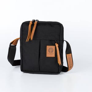 May include: A black canvas crossbody bag with brown leather accents and a zip closure. The bag has a small pocket on the front with a brown leather patch that says "LANDMADE" in black lettering.